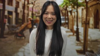 Kalabalık bir sokakta gülümseyen genç bir kadın gölgeli ağaçların altında bulanık şehir geçmişiyle açık havada canlı bir şehir atmosferini yakalıyor..