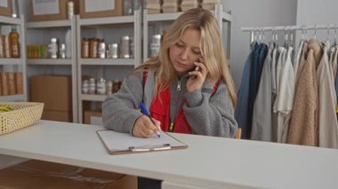 Bir hayır kurumunda gönüllü olarak çalışan bir kadın, içeride bağışlanan eşyalar ve kıyafetlerle telefonda konuşurken bir panoya yazı yazıyor..