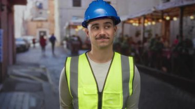Yansıtıcı yelek giyen genç İspanyol bir adam bir restoran ve kahve dükkanının terasında duruyor, iş ve rahatlama karışımı bir şey taşıyor..