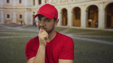 Kırmızı şapkalı ve tişörtlü bir adam üniversite binası avlusunda çeneye dokunuyor; sessiz tefekkür.