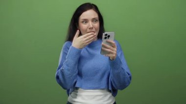 Kumral saçlı bir kadın, yeşil arka planda akıllı telefon kullanırken gülümsüyor, günlük mavi bir kazak giyiyor, modern yaşam tarzında neşeli bir dijital anı yakalıyor..