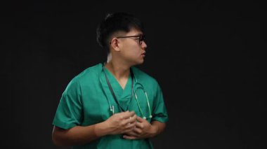 Siyah arka planda tek başına duran yeşil önlüklü genç adam profesyonel ortamda steteskop takıyor..