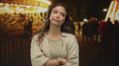 Gece kalabalık bir açık hava fuarında parlak atlıkarınca ışıkları altında çapraz kollu bir kadın; reddedilme.