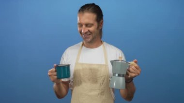 Parlak mavi bir stüdyoda kahve fincanı ve moka demliği tutan önlük giymiş gülümseyen bir adam. Sabah rahatlığı ritüeli..