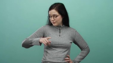 Gözlüklü bir kadın yeşil bir arka plan üzerinde gri bir kazak giyerek, modern gündelik tarzını ve düşünceli ifadesini özetliyor..