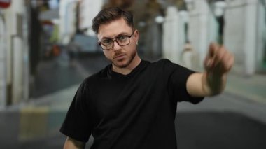 Sakallı ve gözlüklü genç bir adam şehir merkezinde siyah gömlek giyip, tasvip etmediğini belirterek, bir şehir caddesinde küçümseyici bir jest yapıyor..