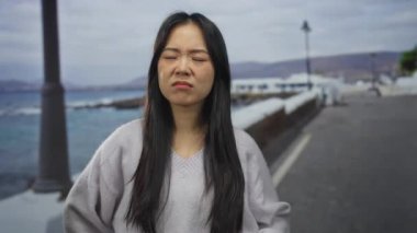 Deniz kenarında gezinen bir kadın bulutlu bir günde dalgın dalgın dalgalanıyor dalgalar çarpıyor, uzun saçlar uçuşuyor ve sokak lambaları manzaralı sahilde geziniyor..