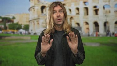 Uzun saçlı sarışın adam Roma 'daki kolezyum binasının yıkıntıları arasında avuçlarını kaldırıyor..