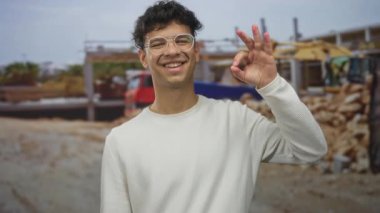 Beyaz süveter giyen ve gözlüklü bir adam inşaat halindeki binanın önünde el yazısı yapıyor. Onaylandı..