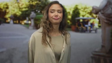 Uzun saçlı bir kadın güneşli yeşil bir parkta dikiliyor, neşeli bir yüz ifadesi sergiliyor, arka planı bulanık ağaçlar ve heykeller çiziyor..