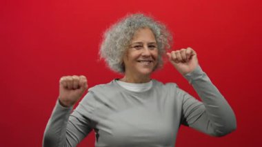 Canlı kırmızı bir duvara karşı neşeyle dans eden kadın, enerjik duruşu ve etkileyici gri saçlı görüntüsüyle mutluluğunu ve güvenini sergiliyor..