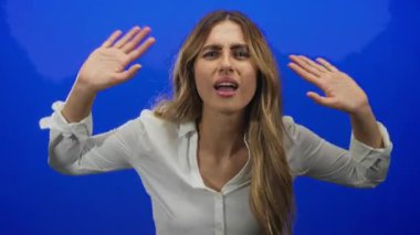 Kadın omuz silkme hareketiyle stüdyoda mavi bir arkaplana el kaldırıyor..