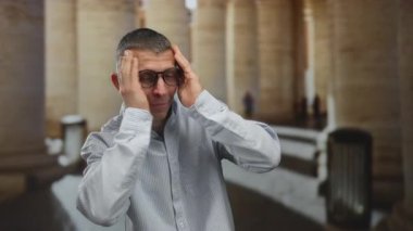 Gözlüklü ve neşeli ifadeli bir adam dışarıda durur, neşeli bir şekilde yüzünü kapatır ve sütunların altında bir şehirde gülümser, şehir ortamında mizah ve duygu gösterisi yapar..