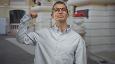 Çizgili gömlekli bir adam arka planda klasik mimarisi olan şehir sokaklarında kaslarını kasıyor, gülümsüyor ve gözlük takıyor..