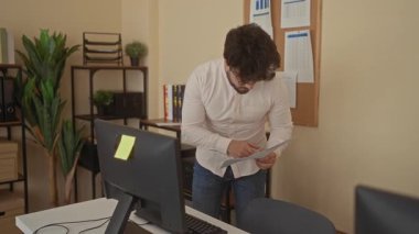 Modern ofiste sakallı genç bir adam haritalar, bilgisayarlar ve bitkilerle çevrili belgeleri inceliyor. Odaklanmış çalışma ortamını gösteriyor..