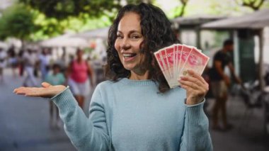 Restoran terasında gülümseyen bir kadın İsrail şekellerini elinde tutarken, bir yandan da paradan söz ediyor ve canlı bir ortamda paranın varlığını vurguluyor..