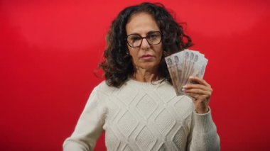 Gözlüklü bir kadın canlı kırmızı bir arka plana karşı Rus rublesi tutarken ciddi ve odaklanmış görünüyor, örgü örülmüş bir kazak giyiyor..