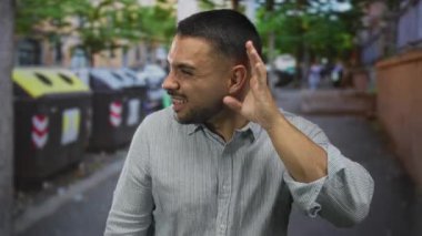 Genç Hispanik adam şehir caddesinde geri dönüşüm konteynırının yanında kulak fincanı; dinleme merakı meydan okuma.