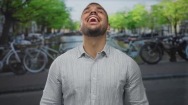 Kırpılmış sakallı, çizgili tişört giyen, kanal ve bisikletlerle gülümseyen elleri yanaklarına bastıran bir adam..