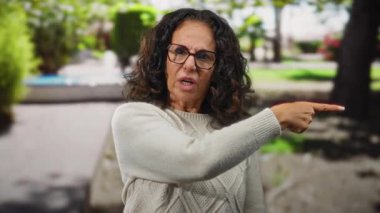 Kadın kızgın bir şekilde park ortamını işaret ediyor. Etrafı yeşillik ve ağaçlarla çevrili. Gökyüzünün altında..