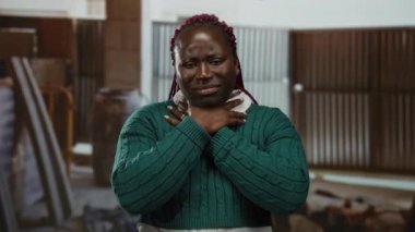 İnşaat alanında boynu ağrıyan, yeşil kazak giyen, iş ortamında endüstriyel aydınlatma altında rahatsızlığını ve yorgunluğunu ifade eden bir kadın..