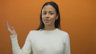 Parlak turuncu arka planda kollarını açmış gülümseyen bir kadın beyaz bir süveter giyiyor.