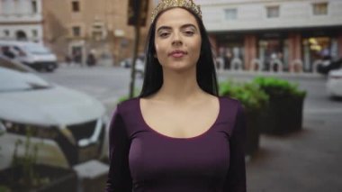 Taçlı İspanyol kadın, açık havada mor bir elbise içinde, kendine güvenerek ayakta duruyor. Gülümseyerek gözüne işaret ediyor..