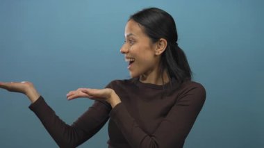 Genç İspanyol kadın, izole edilmiş mavi bir duvara karşı heyecanlı bir şekilde sunum yapıyor. Elleri açık ve neşeli bir ifade sergiliyor..