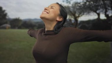 Yeşil bir parkta kollarını açmış ve gülümseyerek doğanın tadını çıkaran bir kadın, dışarıda özgürlük ve mutluluk taşıyor..