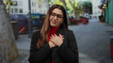 Gözleri kapalı, elleri yüreğinde gülümseyen bir kadın canlı bir şehir caddesinde kırmızı bluz ve siyah ceket giymiş, sıcakkanlı ve kendine güvenen bir kadın..