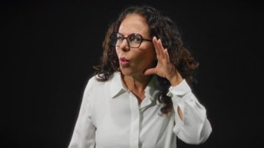 Kıvırcık saçlı ve gözlüklü bir kadın siyah arka planda şaşkınlık yaratıyor, beyaz gömlek giyiyor ve karanlıkta yalnızken el hareketleriyle duygularını ifade ediyor..