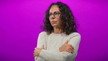 Kollarını kavuşturan kadın canlı mor arka planda soğuk görünüyor. Kıvırcık saçları ve gözlükleriyle rahatsızlık ve zarafet duygusu taşıyor..