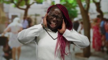 Pembe örgülü ve gözlüklü bir kadın dışarıda bağırıyor, eller kulaklarda, kalabalık bir şehir caddesinde insanlar ve ağaçlarla çevrili, beyaz bir süveter giyiyor..