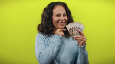 Meksika pezosu tutan orta yaşlı bir kadın sarı arka plana gülümsüyor, para ve kültürel bağlam gösteriyor..