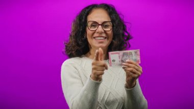 Elinde Endonezya rupiası banknotları tutan bir kadın canlı mor arka planda kameraya özgüven ve pozitiflikle bakıyor neşeli, izole bir ortamda..