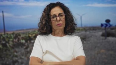 Orta yaşlı İspanyol kadın yel değirmeni tarlasında çıplak kollarını kavuşturuyor..