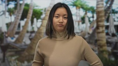 Şehir sokağındaki genç Çinli kadın, bulanık palmiye ağacına karşı ihtiyatlı bir jest yapıyor. Bu da açık hava etkileşimini ve kültürel ifadeyi ima ediyor.