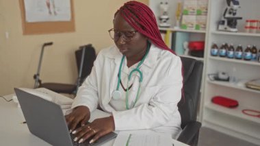Steteskoplu bir kadın klinikte dizüstü bilgisayarla çalışıyor. Arka planda modern ekipmanlarla tıbbi danışma sırasında profesyonel odaklanma gösteriyor..