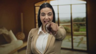 Genç İspanyol kadın gülüyor ve evin içinde büyük pencereleri olan rahat bir oturma odasını işaret ediyor..