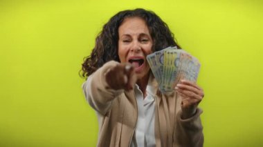 Romanyalı banknotları canlı sarı bir arka planda tutan kadın, neşeli bir ifadeyle para gösterirken enerjik bir şekilde ileriye bakıyor..