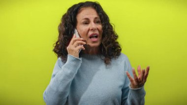 Sarı arka planda telefonda konuşan kıvırcık saçlı bir kadın, el hareketleriyle ifadesini gösteriyor, mavi bir kazak giyiyor, münferit bir yetişkin kadın..