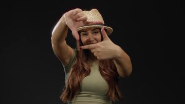 Uzun saçlı, şapka çerçeveli elleri olan genç bir kadın neşeli bir şekilde siyah arka planda izole edilmiş, neşeli ve yaratıcı bir ifade sergiliyor..