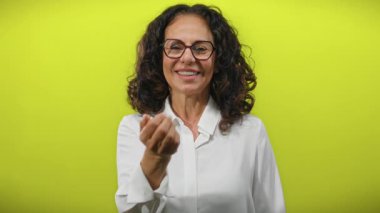 Kıvırcık saçlı İspanyol kadın elini kameraya doğru uzatıyor, canlı sarı bir arka plana doğru, kendine güven ve yakınlık yayıyor..