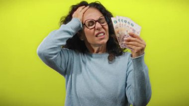 Arjantin banknotları tutan orta yaşlı bir kadın, canlı sarı bir duvara karşı hayal kırıklığını ifade ediyor..