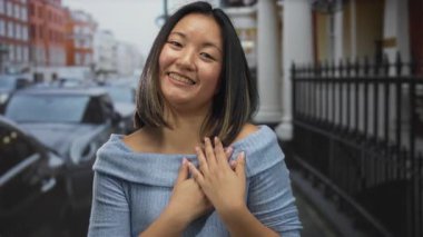 Şehir caddesinde gülümseyen bir kadın mavi süveter giyiyor. Arka planda arabalar ve binalar var. Şehir ortamında mutluluk ve güveni ifade ediyor..