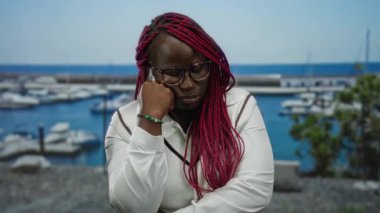 Gözlüklü ve kırmızı örgülü bir kadın, arka planda tekne ve deniz olan bir limanın yanında düşünceli bir şekilde oturur, berrak mavi gökyüzünün altında derin bir düşünce ve dinginlik hissi yayar..