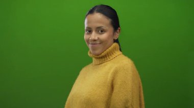 Sarı kazaklı bir kadın, yeşil arka plana parmak atarak buraya geliyor, özgüvenli ve davetkar bir şekilde gülümsüyor, sıcaklığı ve yakınlığı gösteriyor..