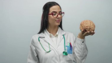 Steteskoplu İspanyol kadın doktor, arka planda beyaz bir duvar karşısında bir beyin modeli tutuyor..