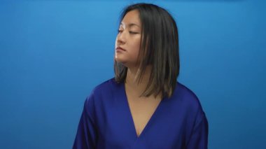 Mavi elbiseli genç Çinli kadın mavi arka planda dikiliyor ve ciddi bir ifadeyle ileriye bakıyor..