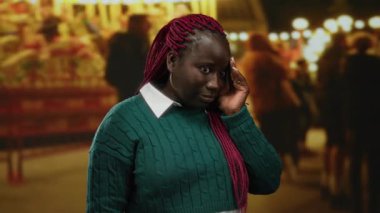 Kırmızı örgülü, yeşil kazaklı bir kadın, açık hava panayırında duruyor, bulanık ışıklar ve insanlarla çevrili, konuşurken şaşırmış görünüyor..